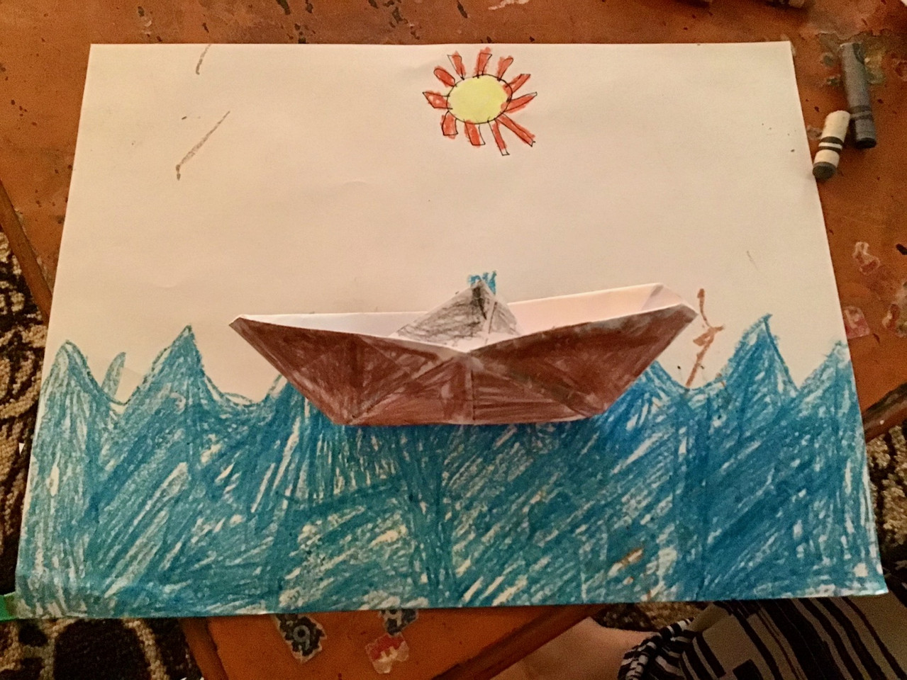 Origami boat colored brown on a drawing of water with a yellow sun