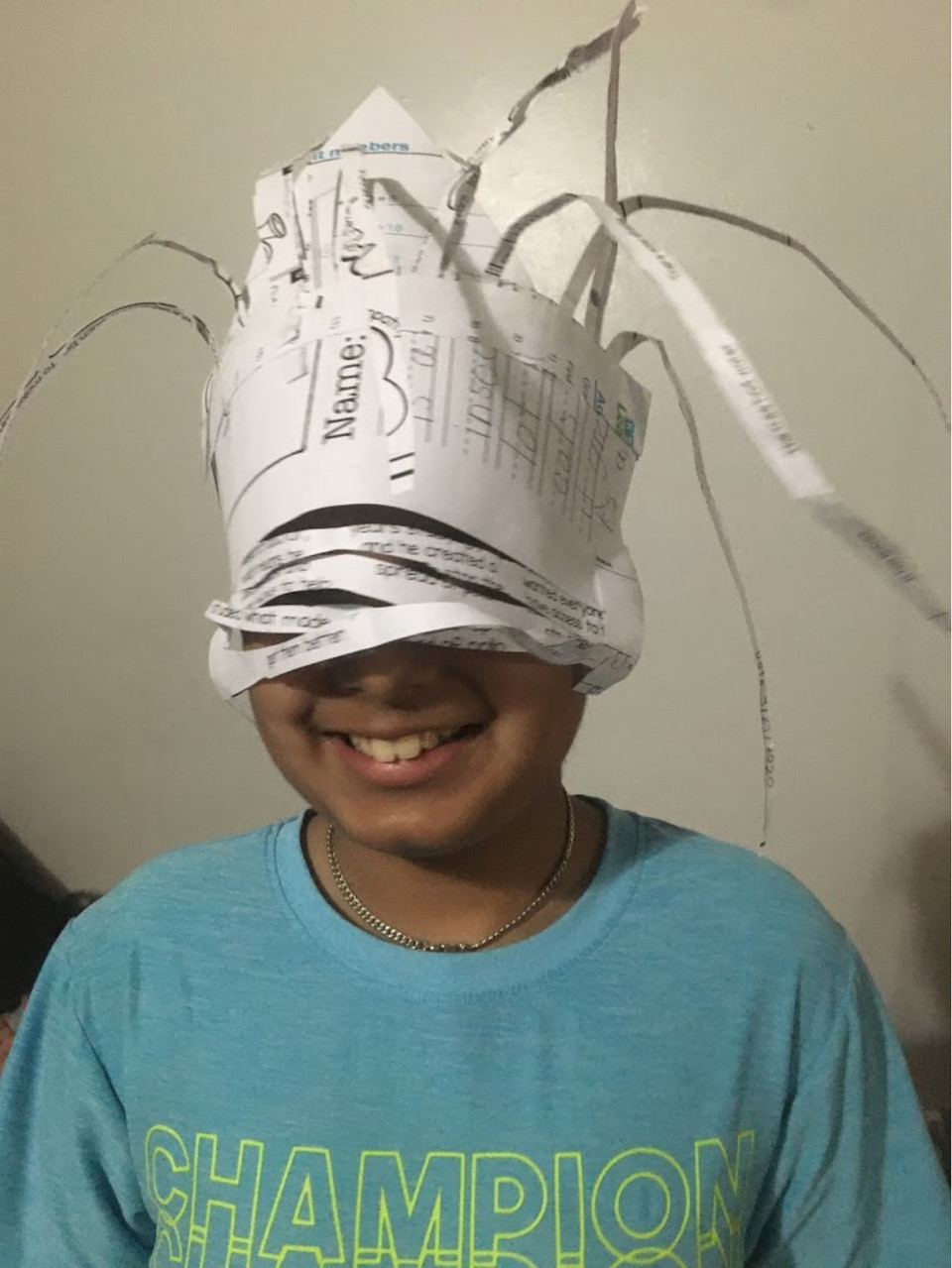 Child in teal shirt wearing a hat of white paper with fronds and cut strips covering their face
