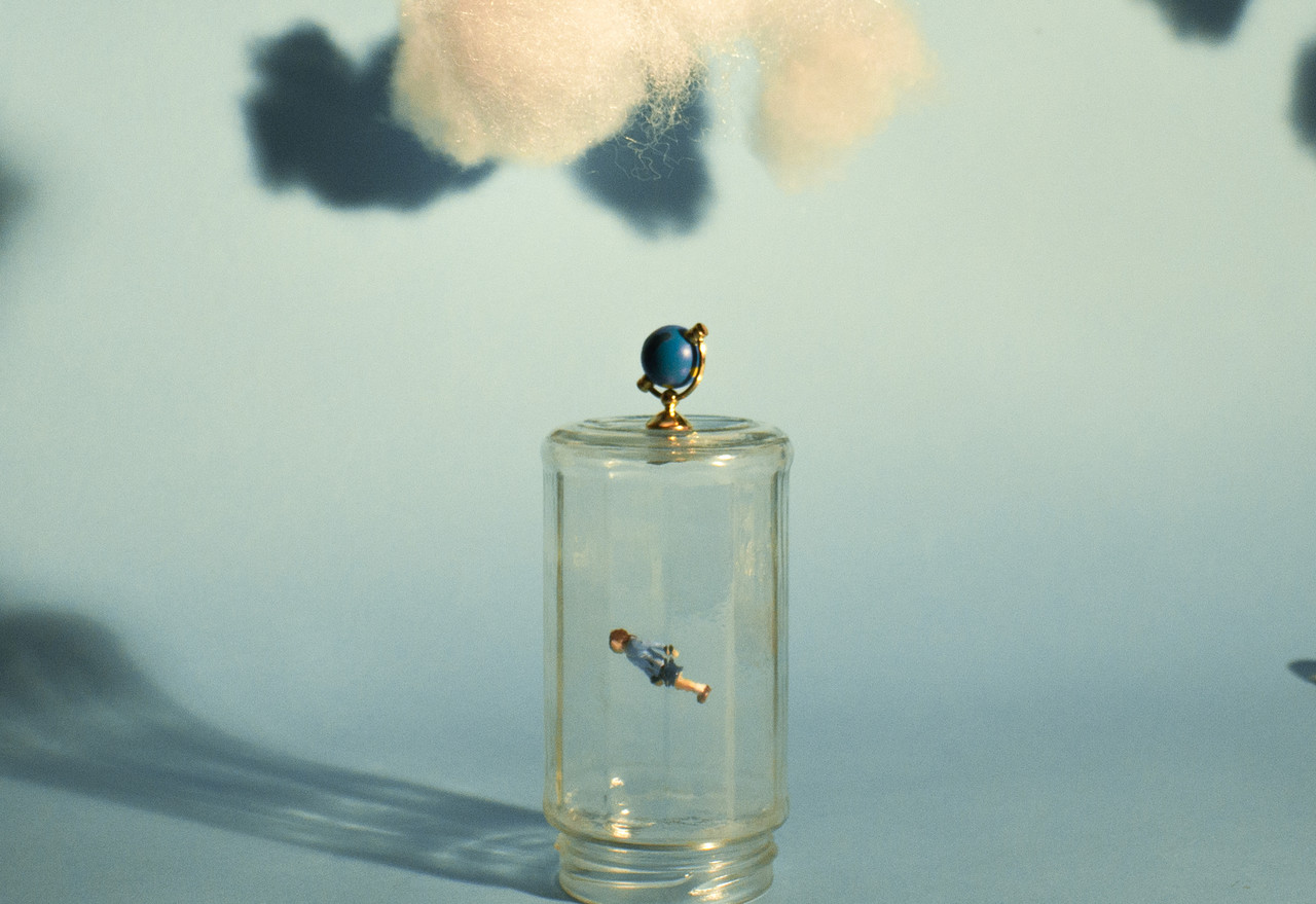 A floating figurine trapped inside a glass mason jar with fake clouds overhead and a baby blue background