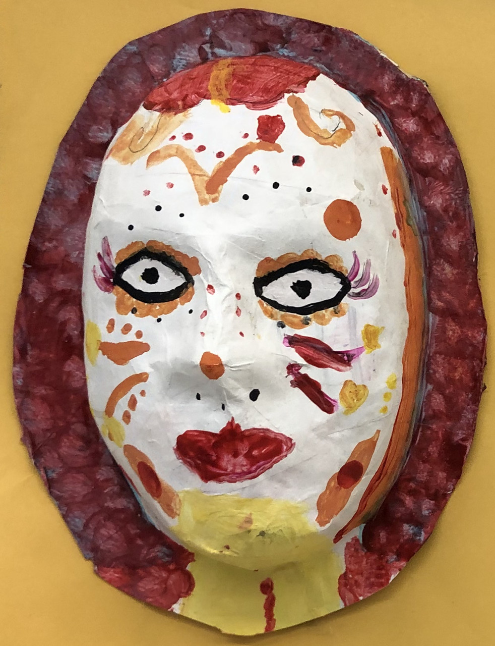 Mask with white background, embellishments around the face of swirls, spots, and lines in yellows, browns, and reds