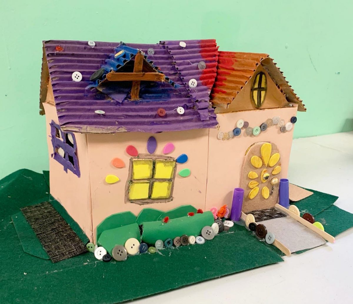 Light pink house with corrugated cardboard roof painted purple, covered with buttons
