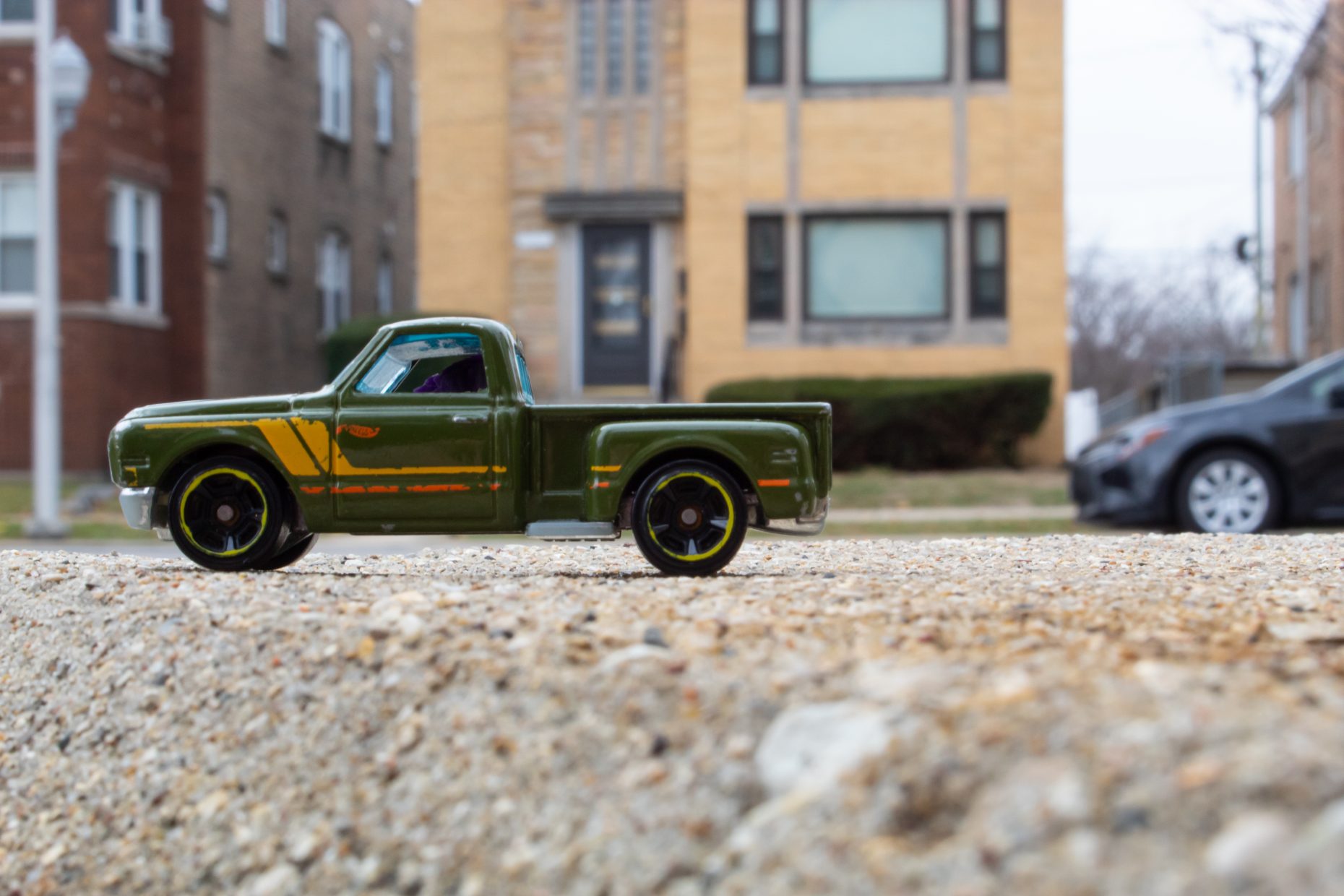Photograph: in focus is a green toy truck on a road. In the background is a brick apartment building. Perspective is manipulated to give illusion the toy car is actually to scale of a real car.
