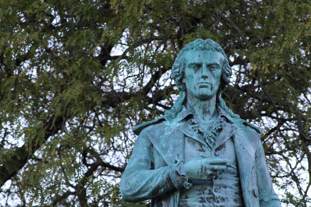 Photo: top half of a stone sculpture of a person holding a pencil with their right hand at chest. Photo is taken outside; tree branches in the background.