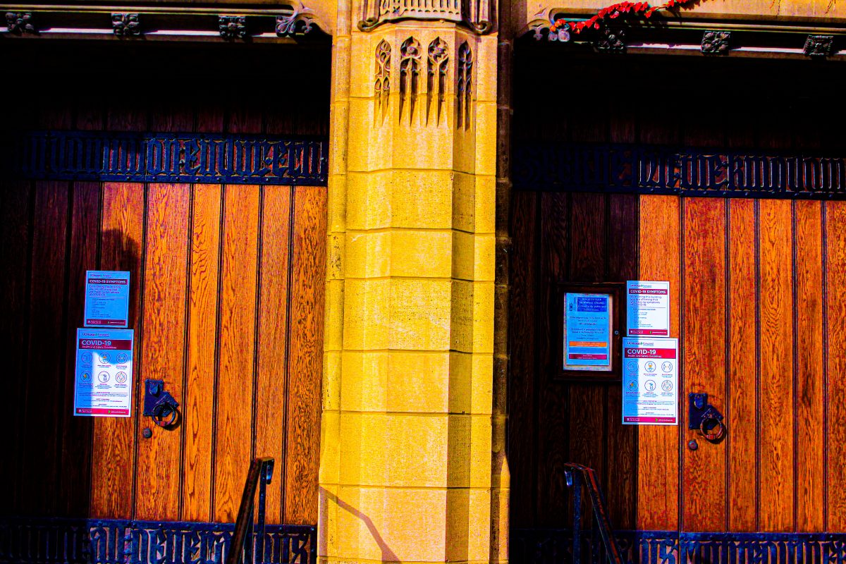 Photograph: Two large wooden doors separated by a stone column. On each door is signage with health guidance regarding COVID 19.