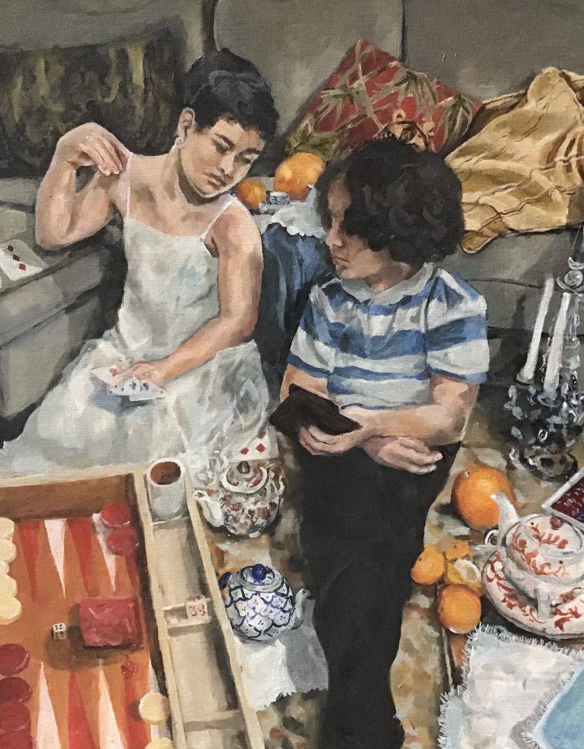 Two people sitting on the ground and leaning against a couch in a domestic space. Person on the left is wearing a white dress and person on the right is wearing black pants and a blue and white stripped shirt. There are teapots, teacups, oranges, candles, and a board game on the floor.