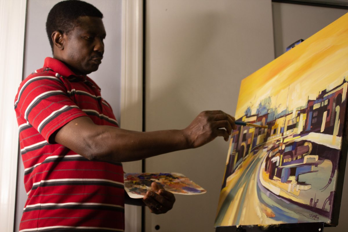 Photograph of a person painting. They wear a red, white and grey stripped polo shirt. They hold a paint palette in their left hand and a paint brush in their right up to the canvas. The painting is of a street scene.