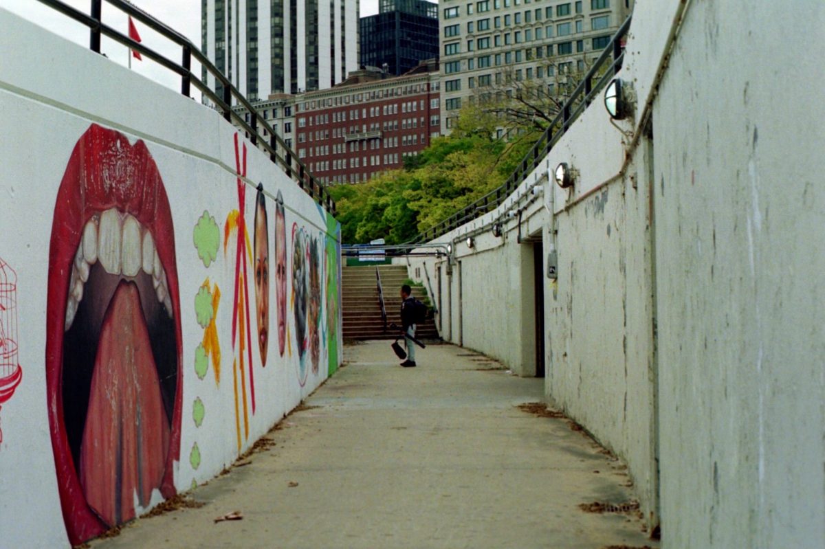 Photograph of subterranean, concrete pedestrian passageway with mural art on left wall of an open mouth and people's faces. A person stands at the distant centered background in front of a flight of stairs.