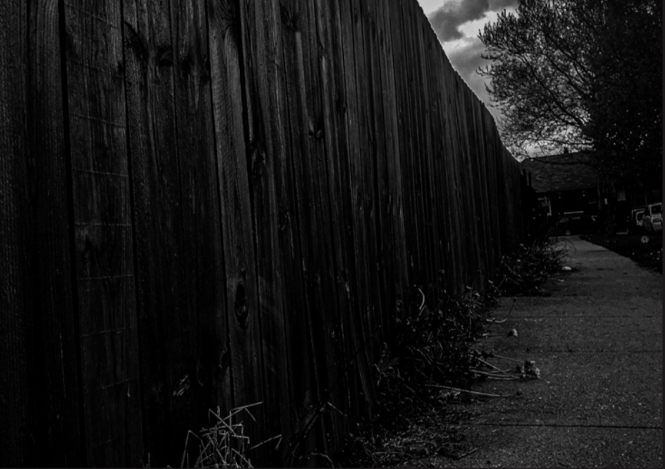 Oblique-view photo of a wooden fence down a street, in dark, night-time grayscale.