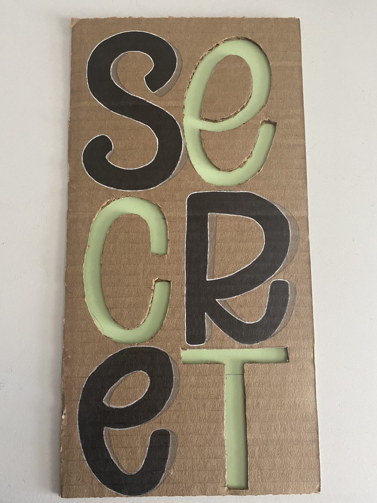 "Secret" on cardbord, with the S, R, and E drawn in black and the E, C, and T cut out and overlayed on a green background