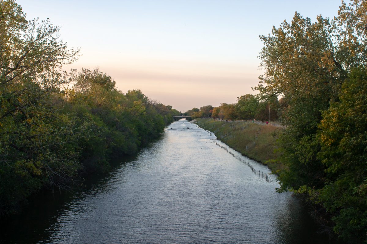 Sunset over the North Branch of the Chicago River, surrounded with trees and natural plants. Two kayakers are on the river, with people walking on adjacent path.