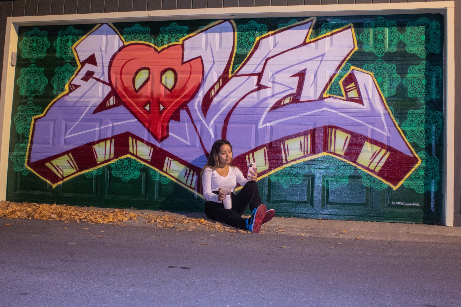 Person sitting with phone and iced coffee in front of a garage with style writing on it.