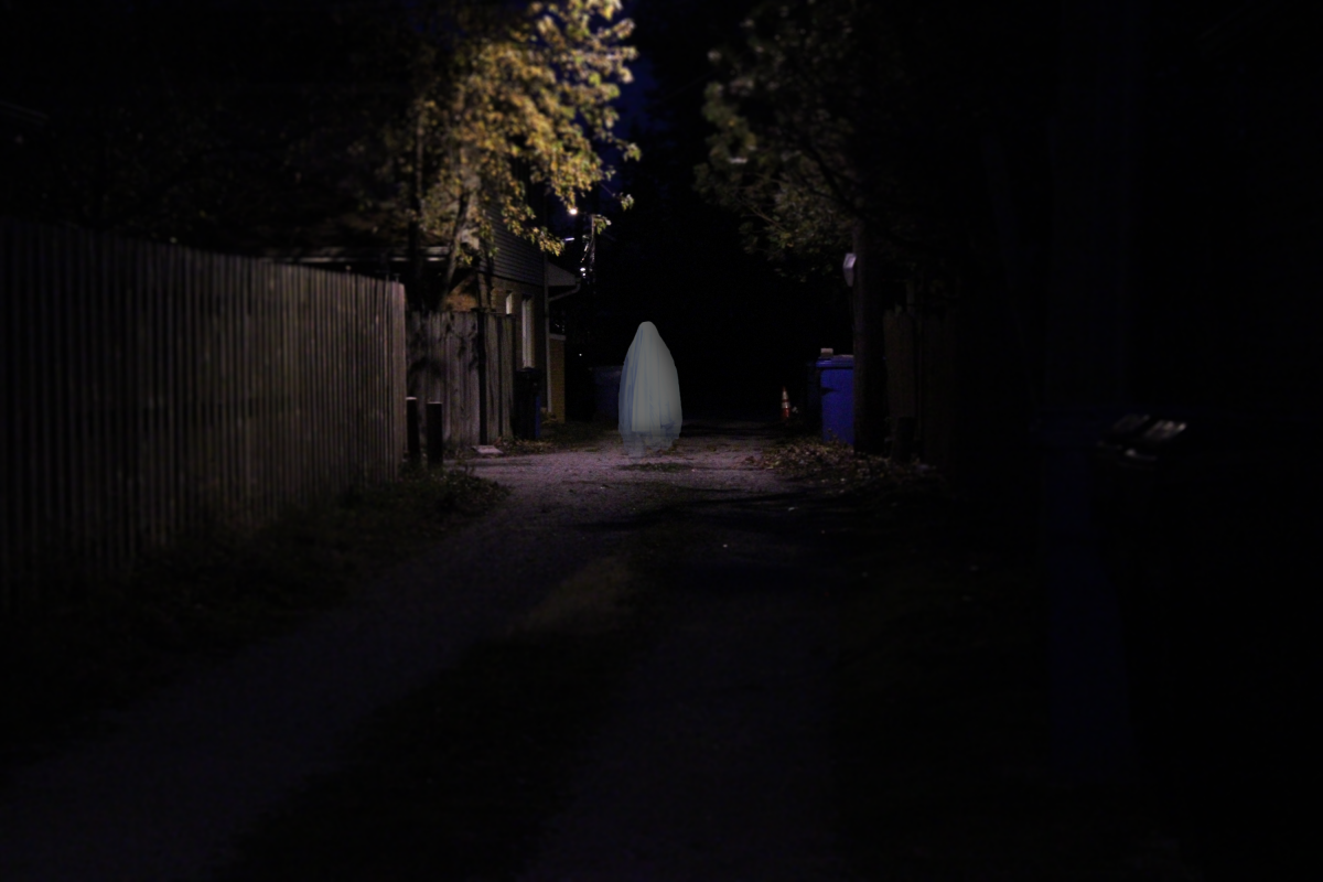 Photo of an alley behind houses at night. A light illuminates a ghostly figure in a white sheet.