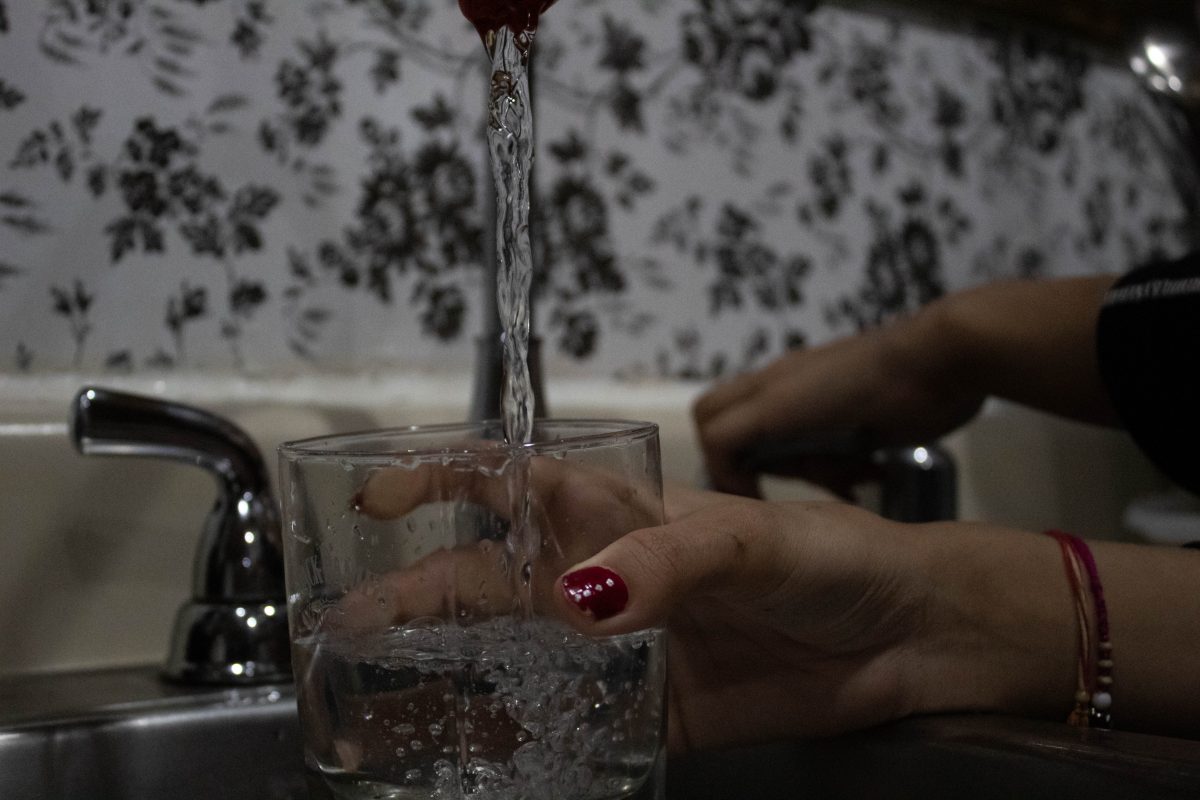 Photograph of water running into a glass from a sink. There is out of focus black and white floral wallpaper in the background.