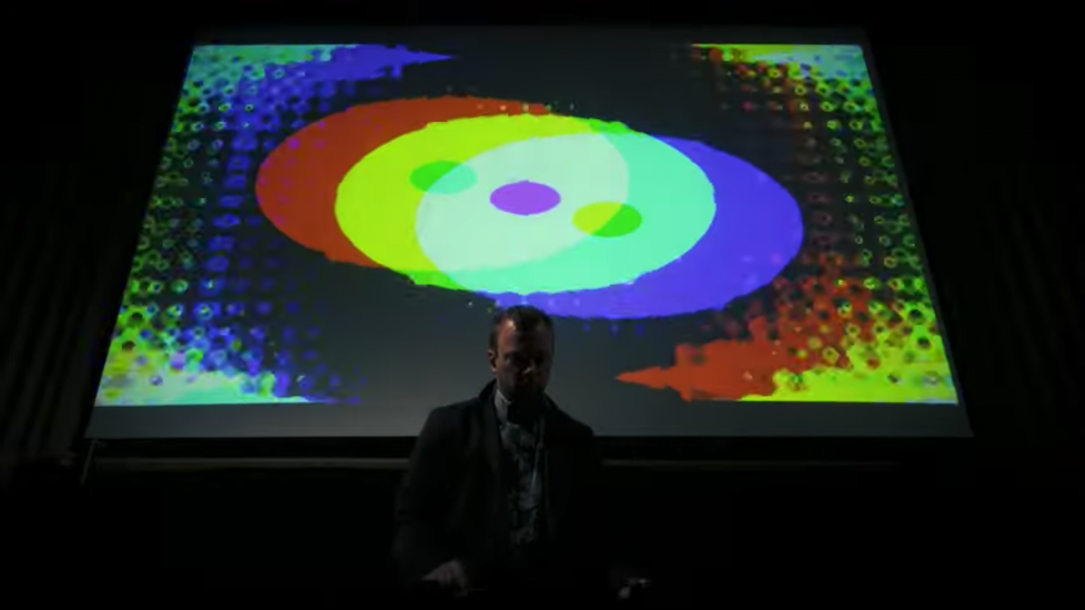 Person in suit playing a mallet instrument. They stand in front of a screen with digital shapes projected on it.
