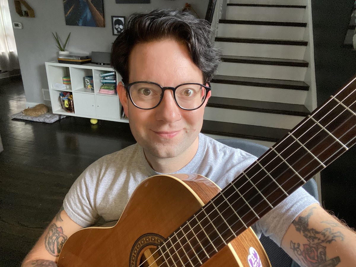 Person with dark hair, glasses, nose ring, and tattoos sits with a guitar in front of a staircase.