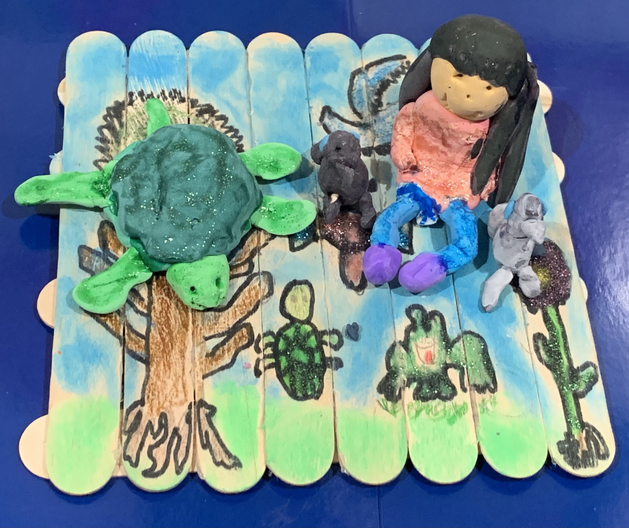 A sculpture of a popsicle stick raft with a girl and turtle made of clay sitting on the raft.