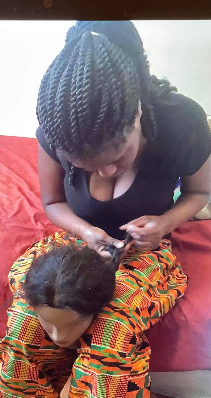 Photograph of a Black person braiding the hair of a large doll in her lap.