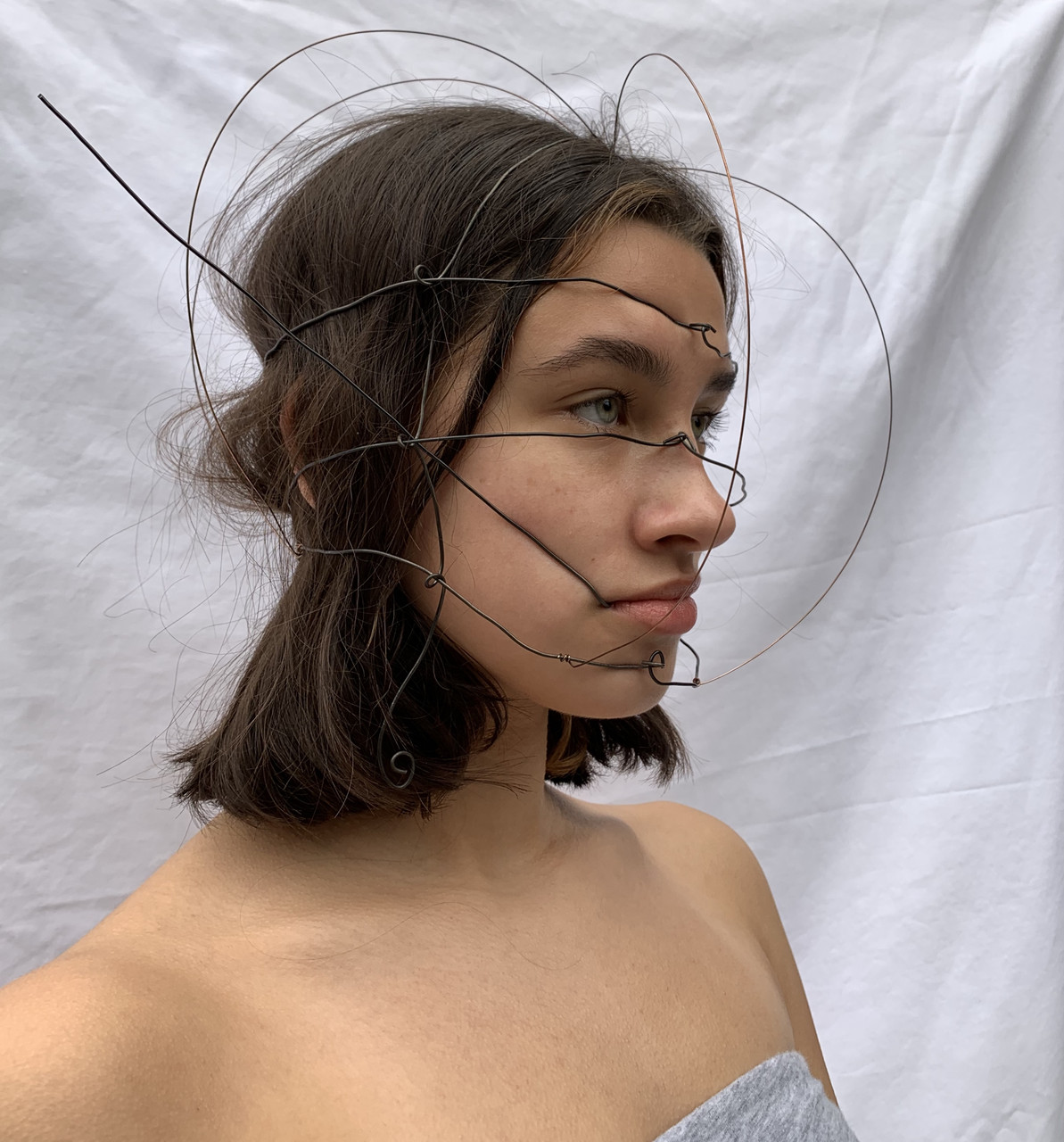 Wire sculpture worn on person's head. The wire loops around and encloses the model's face.
