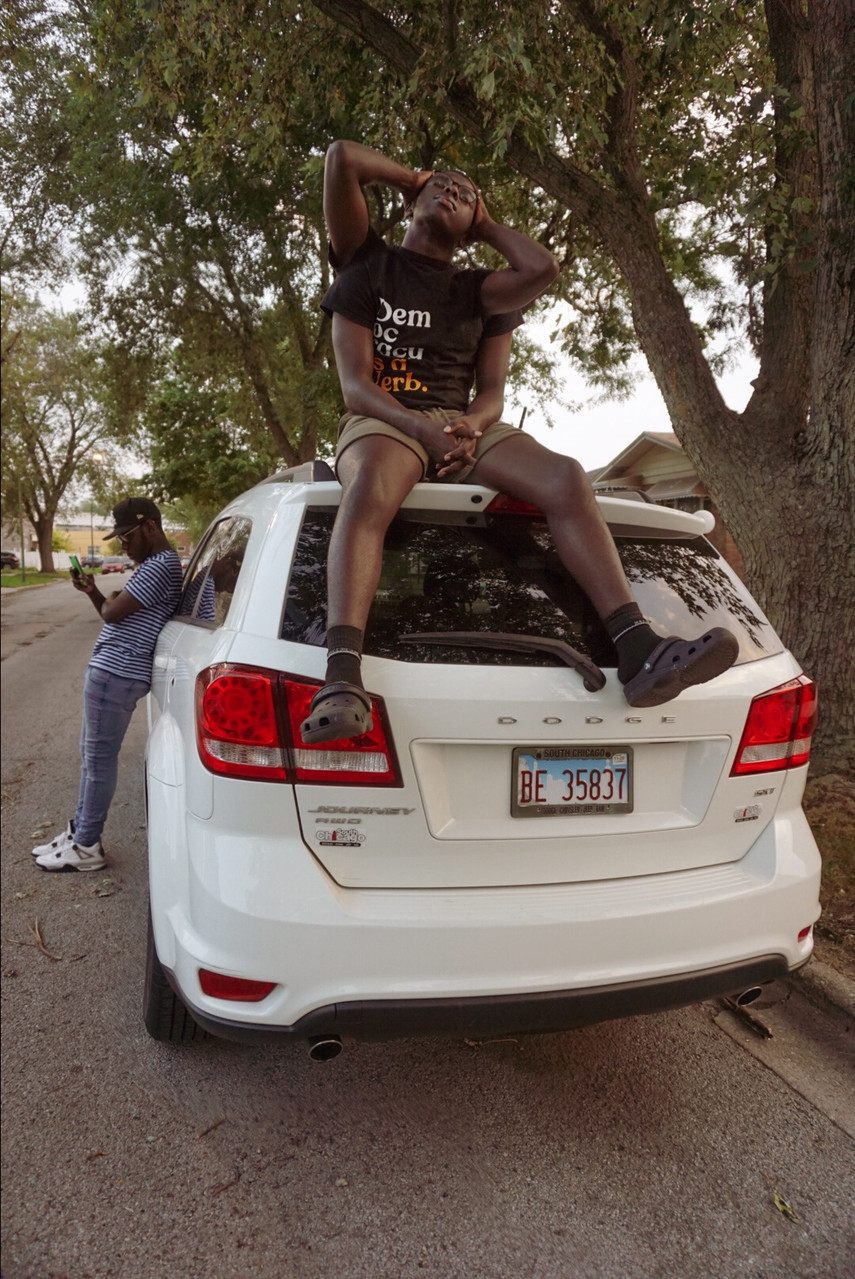 Black person sitting on the roof of a white car, with another Black person leaning against the driver side door.