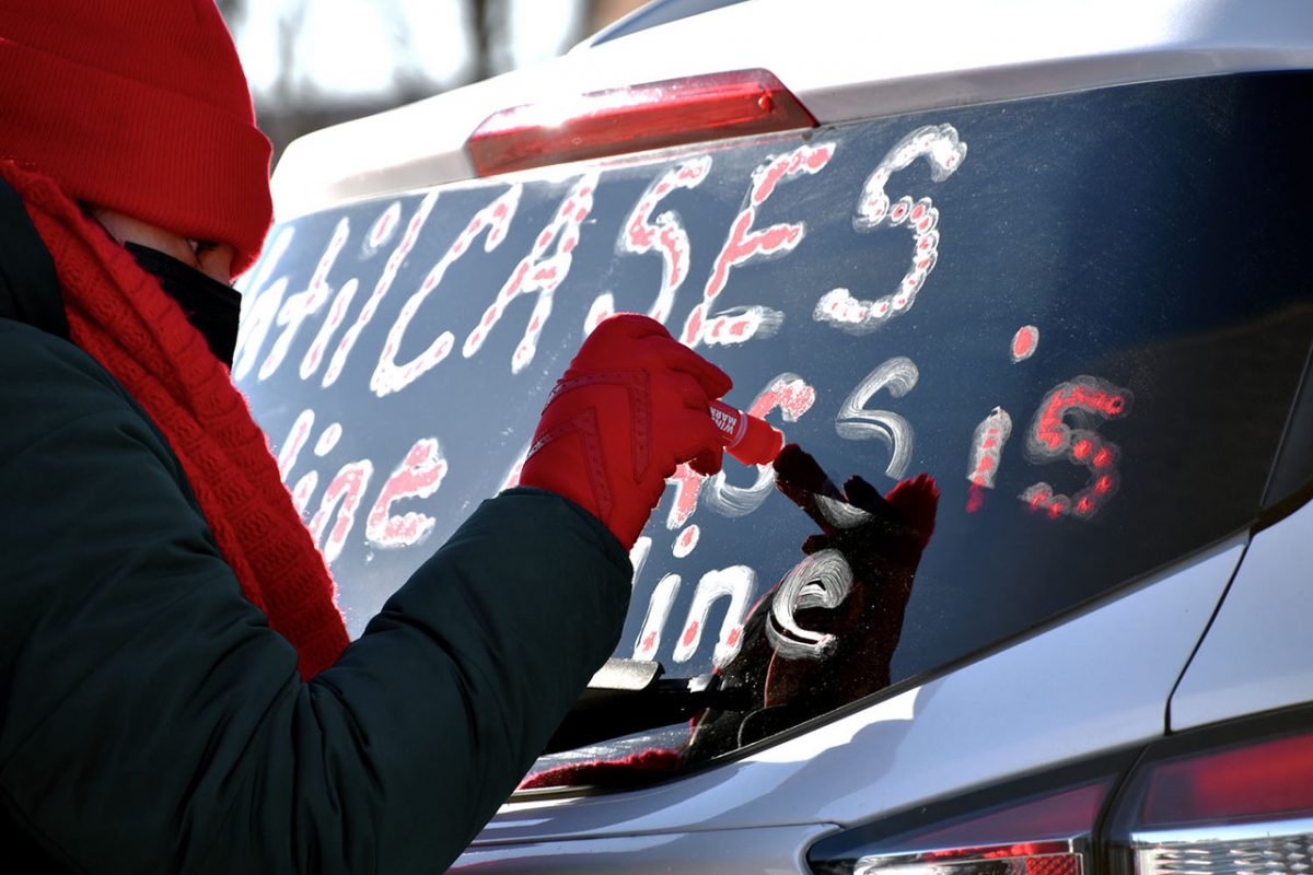 Photo of person in winter gear with a red hat, scarf, & glovesdrawing on the rear window of a car with marker
