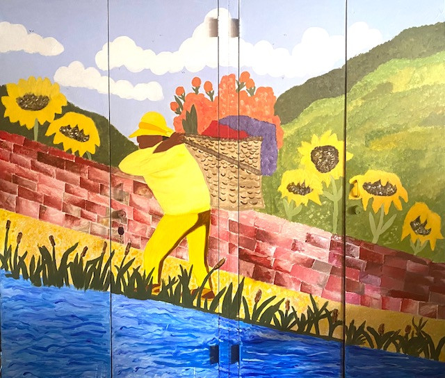 Mural with figure in yellow in front of mountains, next to a brick wall with large sunflowers & water