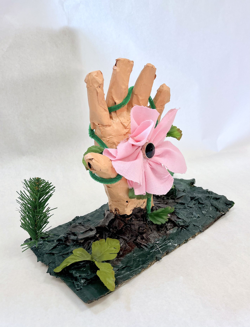 sculpture of pink hand rising from green vegetation, a pink flower comes from the palm