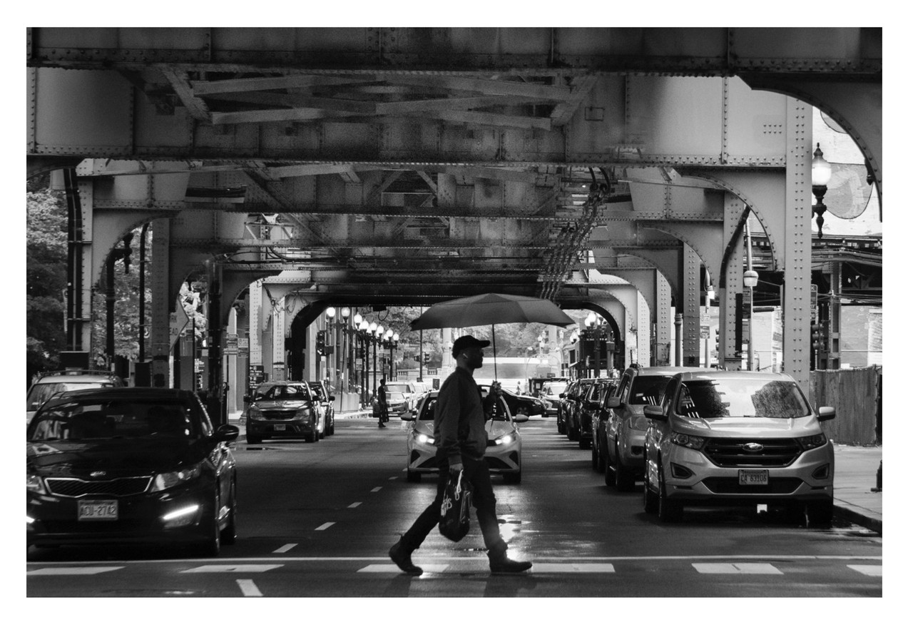 Black and white photograph of a man crossing the street. He walks under train tracks with cars waiting to cross the road next to him.