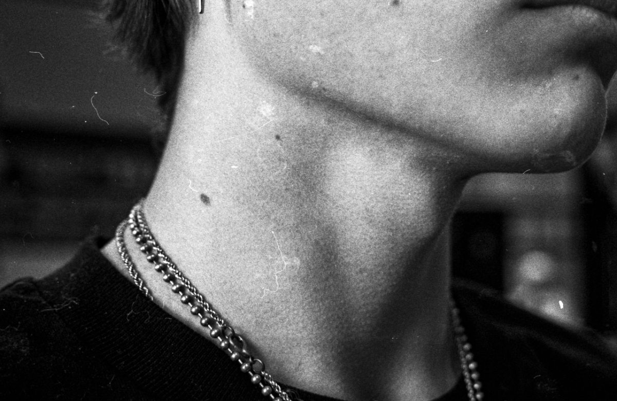 Black and white photograph of a subject's neck, including multiple chain necklaces and a t-shirt.