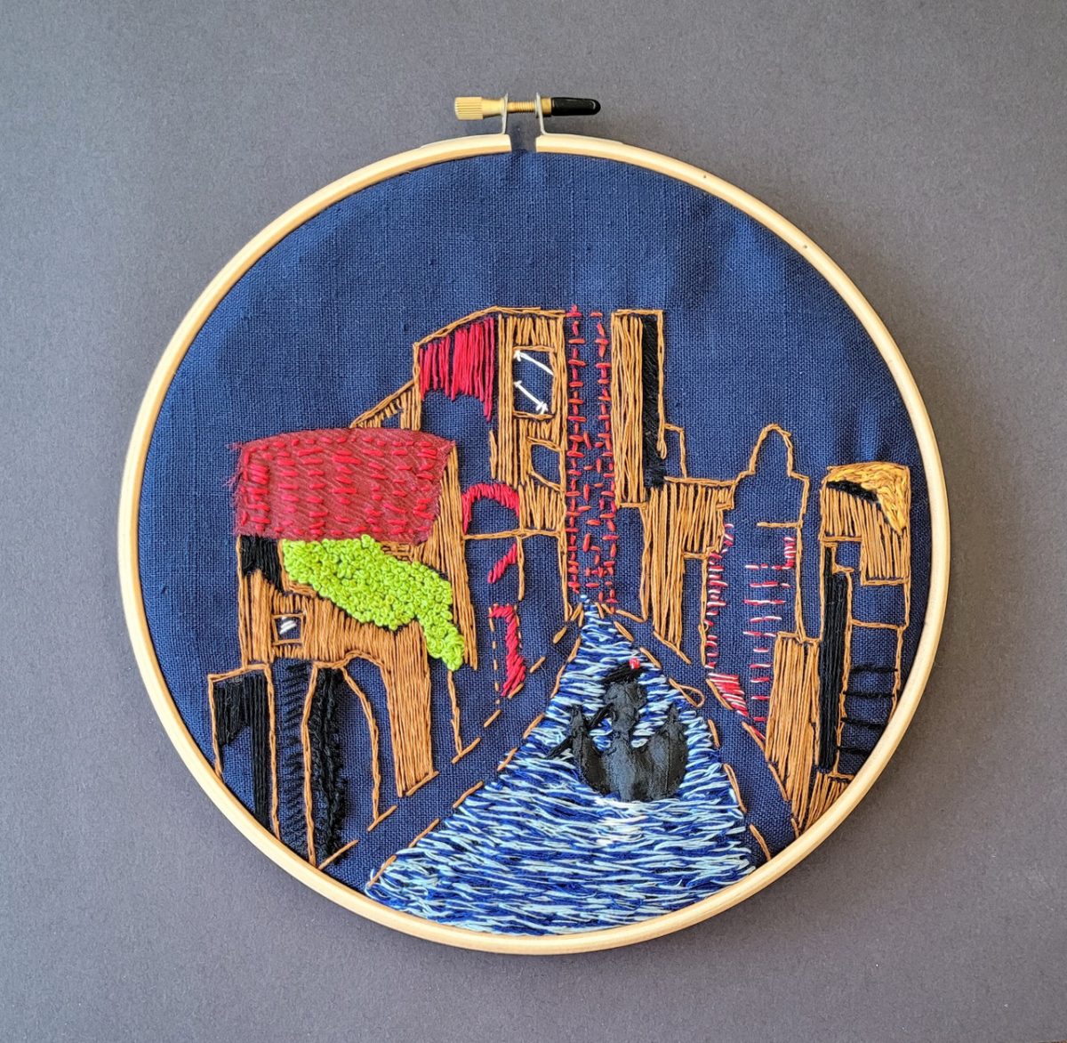 Scene from Venice embroidered on blue fabric. Orange and red buildings line a blue canal on either side with a boat in the center.