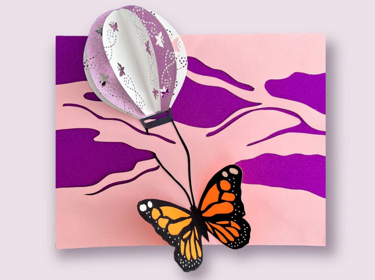 Made of paper construction. A monarch butterfly is carried by a purple and white hot air balloon over a pink and white background page.