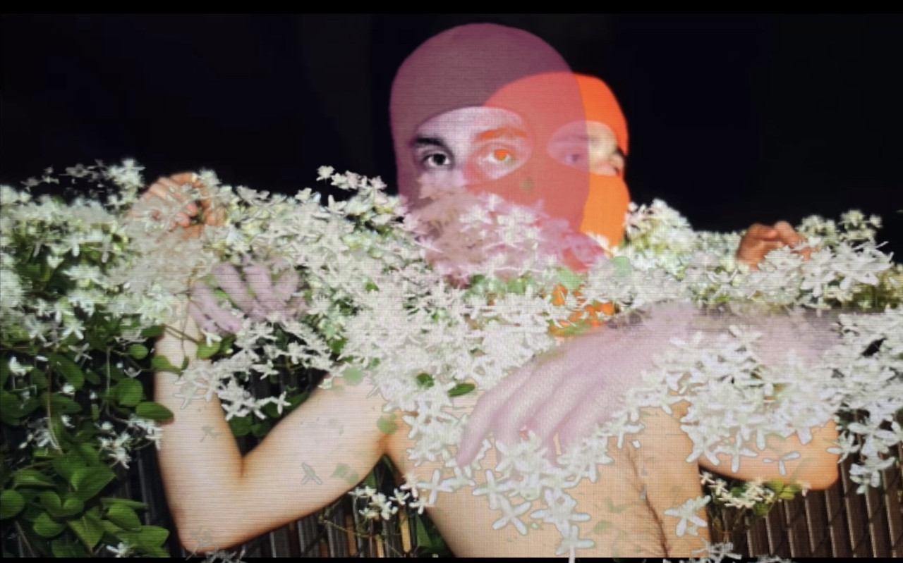 Photograph of what appears to be two men in masks. One holds the other across the shoulder and white flowers pass over both of them.