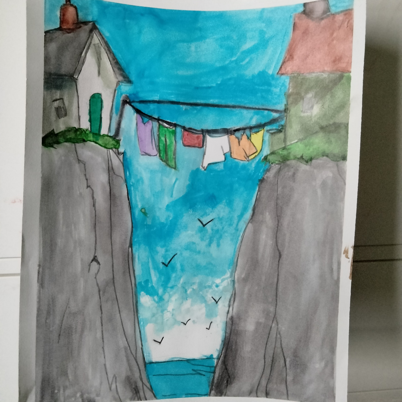 Laundry hangs on a line between two houses which sit on opposite sides of a body of water on a cliffs.