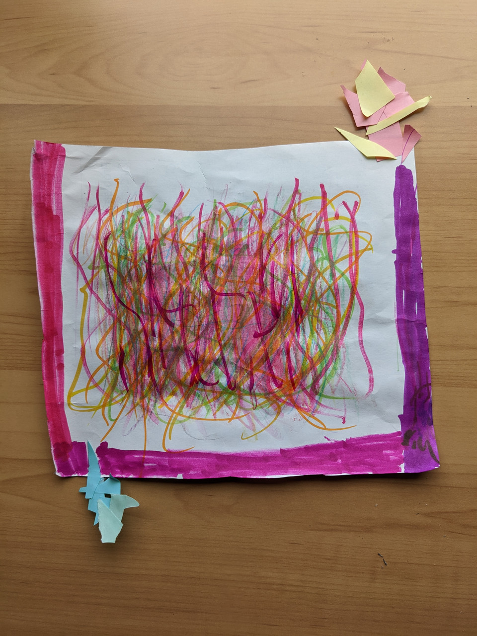 Scribbles of color on white paper are surrounded by a pink and purple outline with construction paper cutouts attached at the corners.