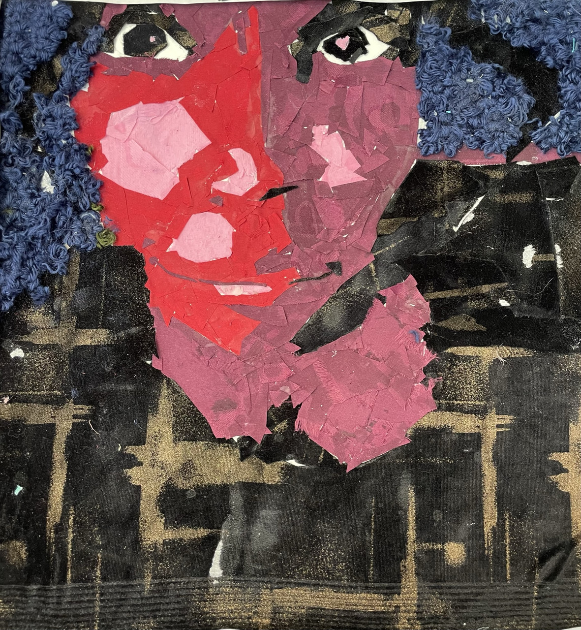 A fabric on board self portrait. The face is pink and red surrounded with blue and black.