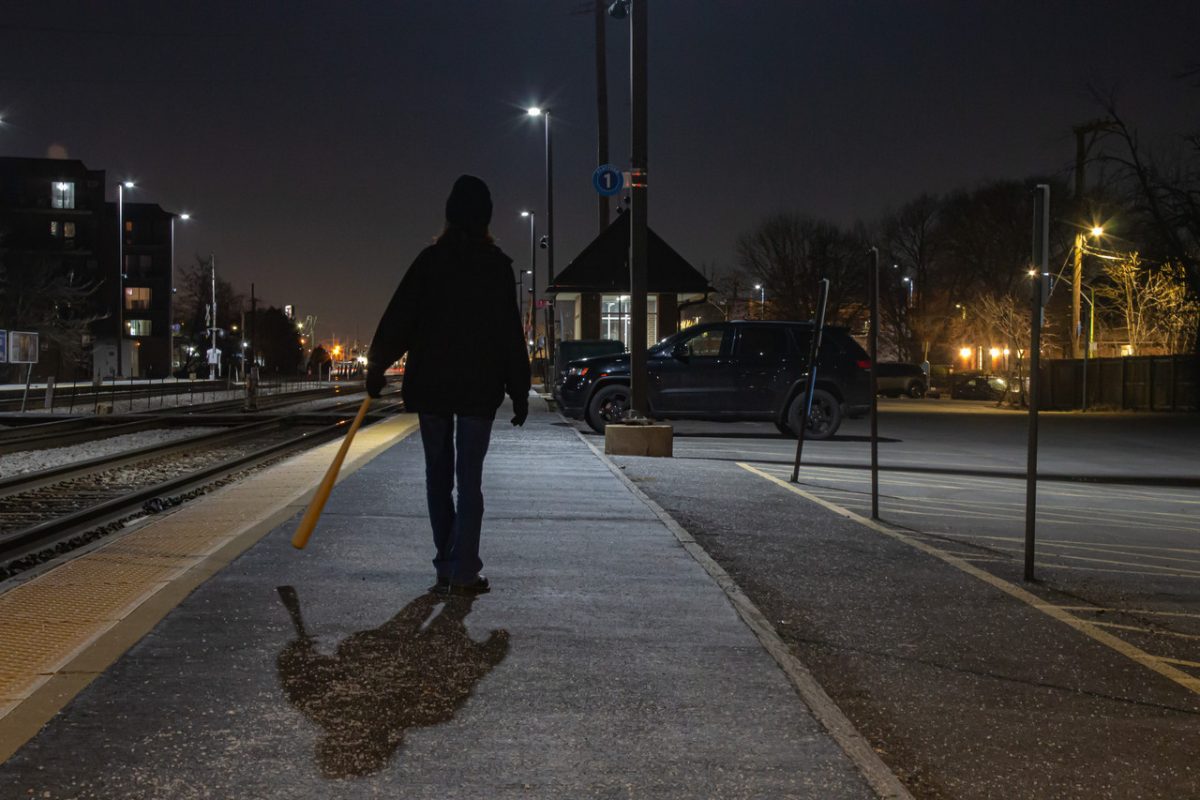 A photograph of a person holding a bat at night walking away from the viewer on a road.