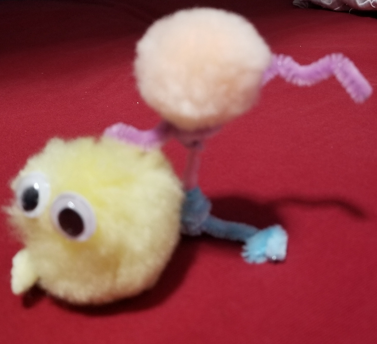 A yellow pom pom with two googly eyes makes a head and is attached to a pink pom pom ( the body) via pipe cleaners.