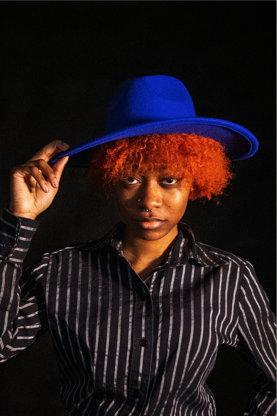 photograph of a young person emerging from a black background with red curly hair, wearing a blue hat