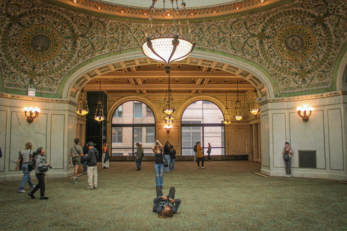 Wide, symmetrical view of a room in the Chicago Cultural Center, with a photographer in the center on the ground facing up, taking a photo.