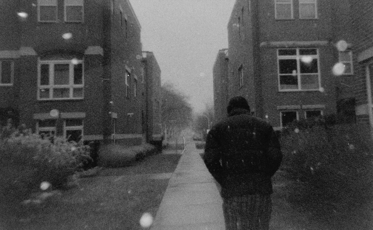 A black and white photo of a person walking on a street between buildings on a snowy day.