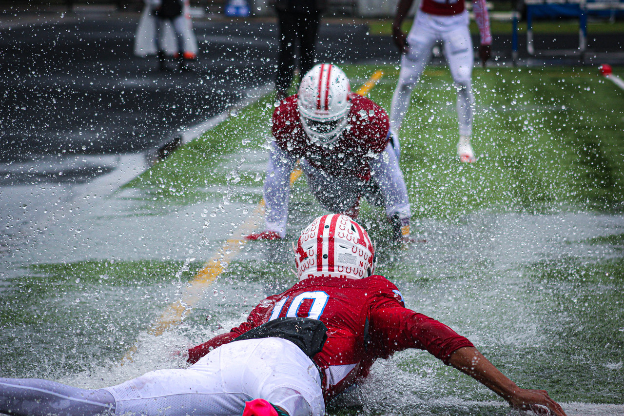 Photo of two football players in red sliding toward each other on wet field. Lots of water comes out of the field.