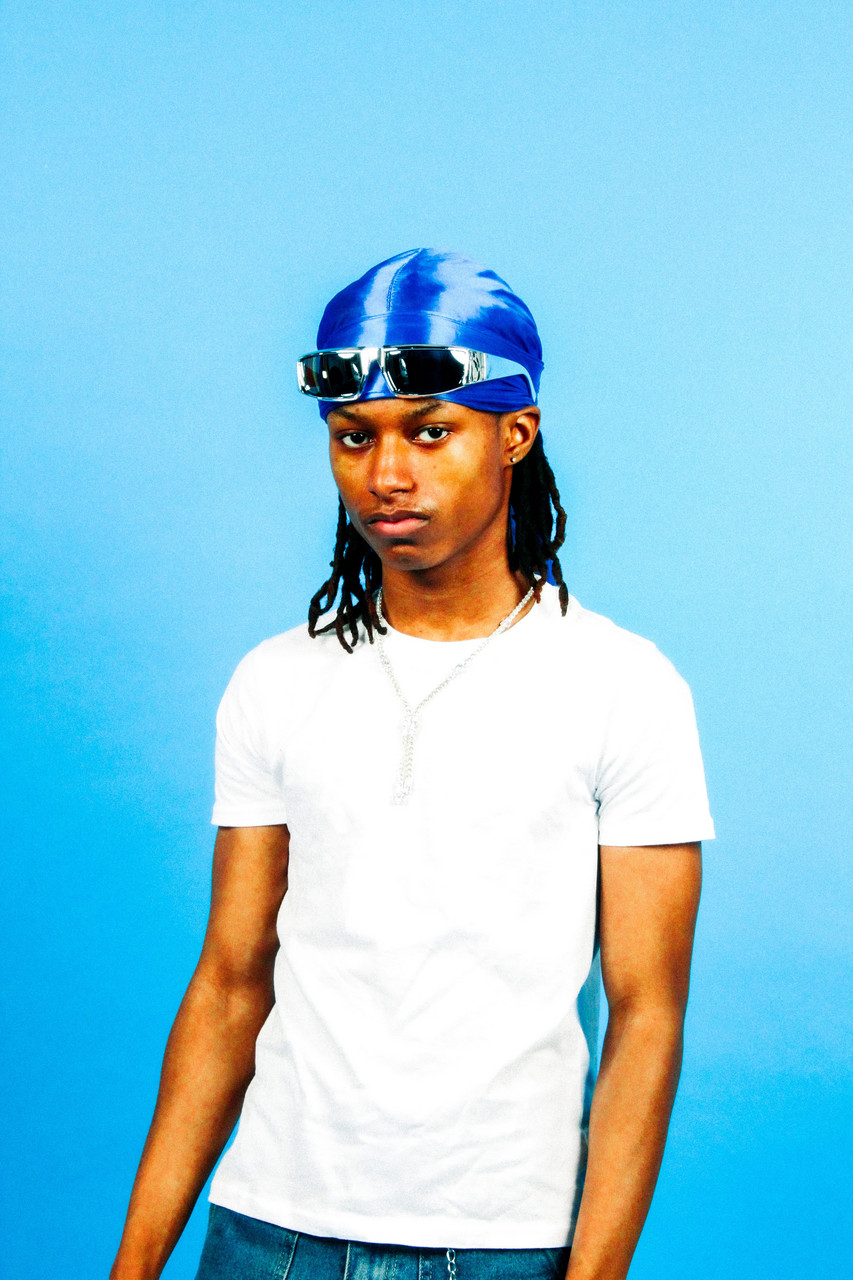 Young man in white shirt with blue head covering stands in front of a blue background.