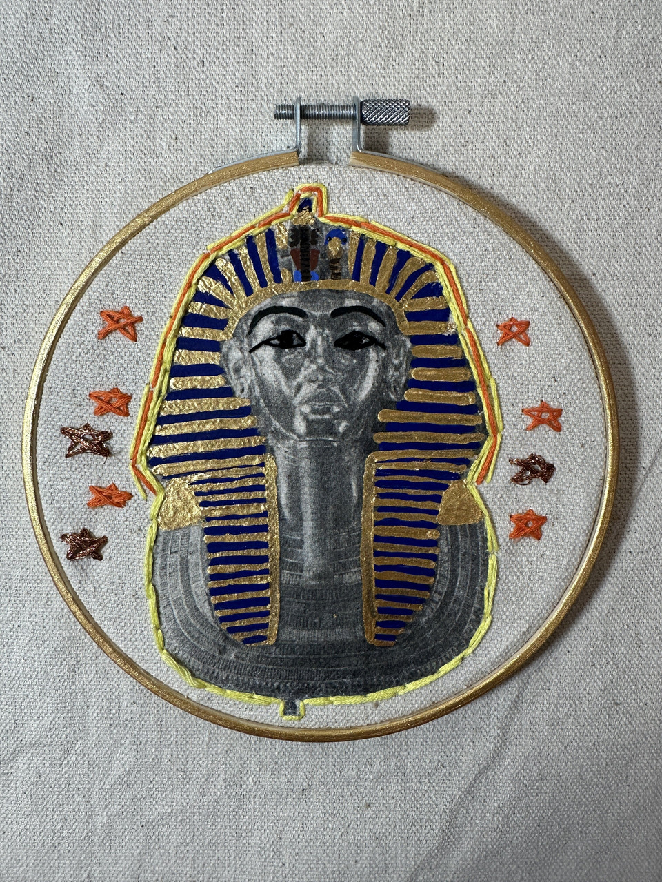The sarcophagus of King Tut painted onto a canvas with an embroidered yellow and orange border. Orange and copper embroider stars surround the portrait