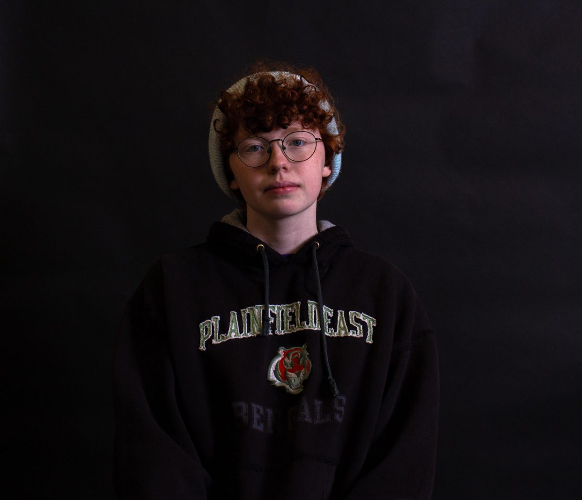 Person staring straight into the camera against a black background. They have red curly hair, blue eyes, pale skin
