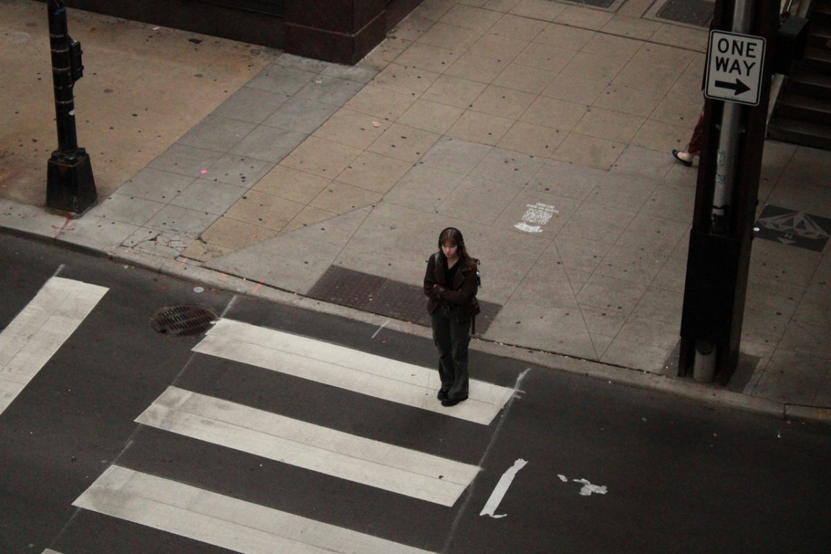 A girl stands at the beginning of a crosswalk. The viewer looks down on the dim street which is otherwise empty.