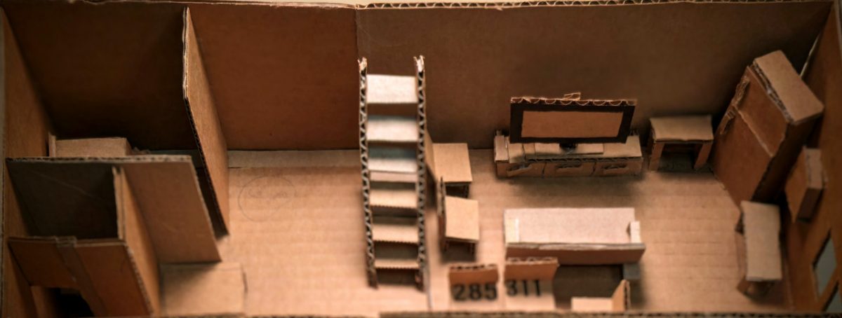 Cardboard sculpture of a room with stairs, a desk with computer, chairs, tables, and a closet.