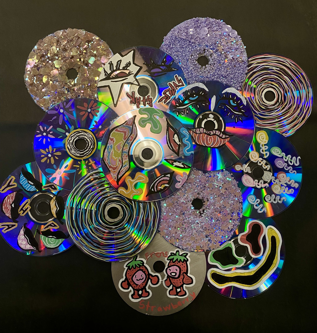 Several CDs, each with unique designs, are layered on top of each other. Several are sparkly and luminescent.