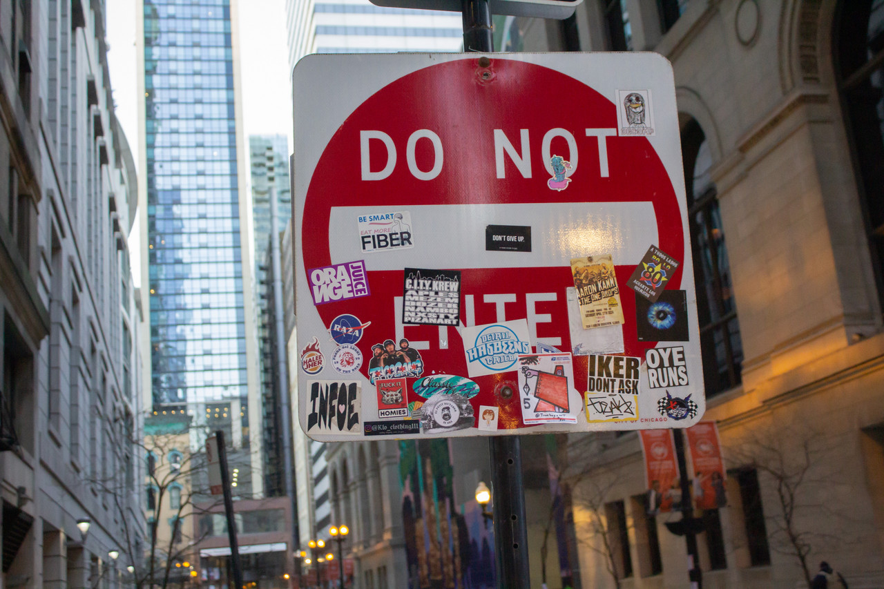 photograph of do not enter sign covered in stickers with city buildings in background