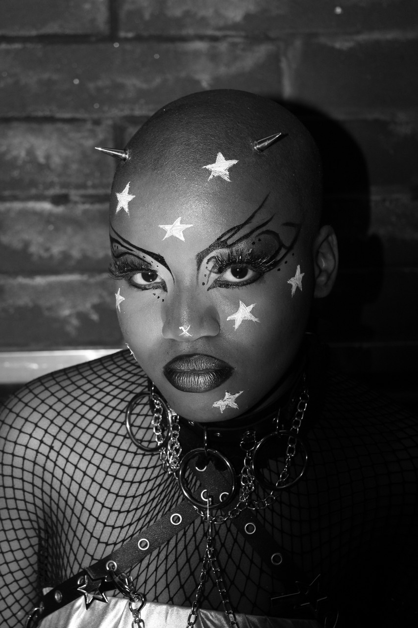 black and white photograph of dark skinned person with dramatic black makeup and white stars on face looking intensely into the camera