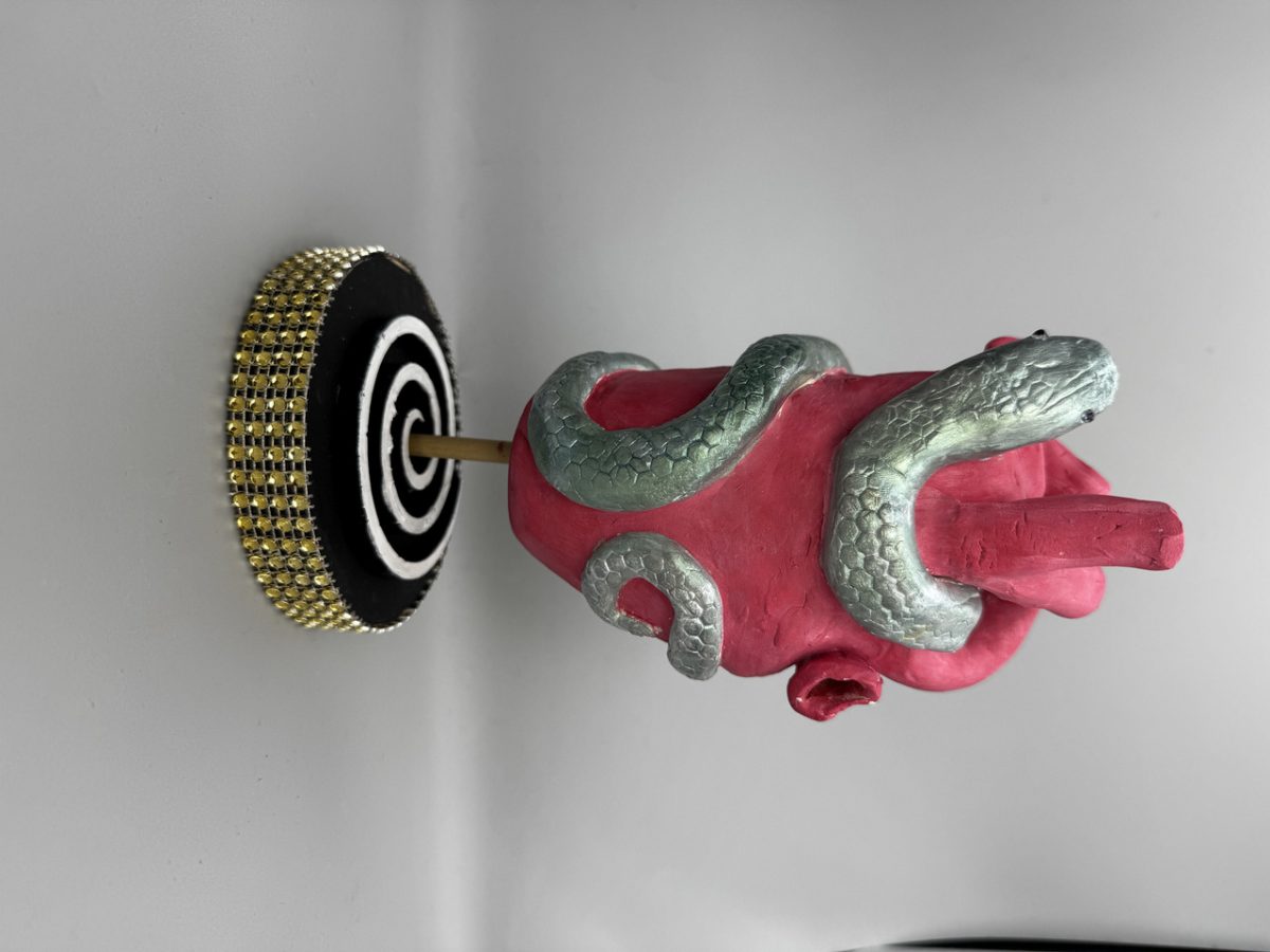 Clay sculpture of anatomical heart with snake wrapped aroun it being held up by stand covered in gold gems
