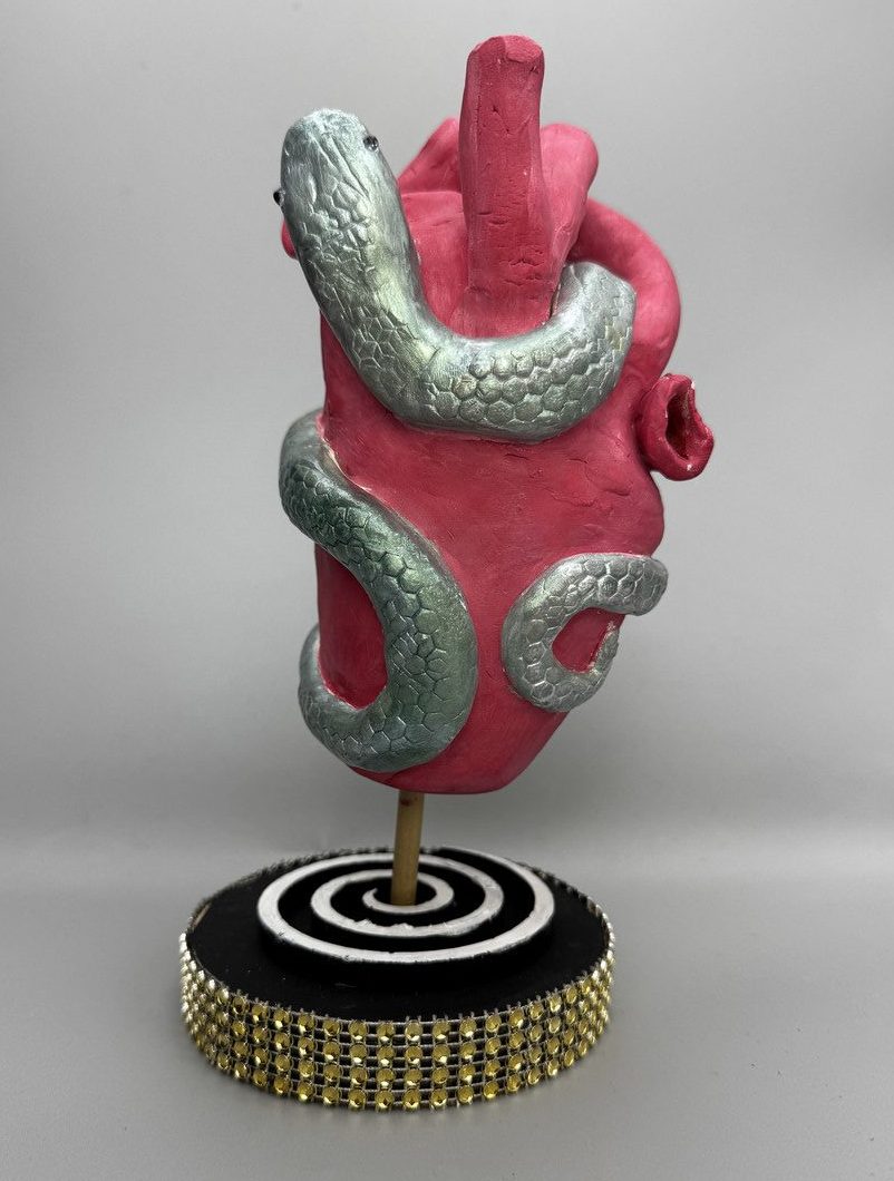 Clay sculpture of anatomical heart with snake wrapped aroun it being held up by stand covered in gold gems